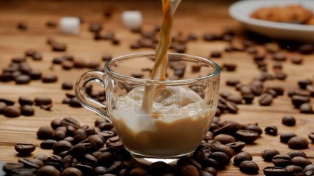 Photo for Streams of coffee and milk pour into a glass cup, filling it with an aromatic drink. Close up of a cup of coffee or milk tea on the kitchen table near coffee beans. Morning ritual, breakfast concept - Royalty Free Image