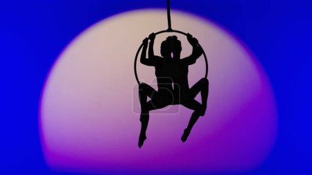Téléchargez les photos : Chorégraphie moderne et acrobaties concept publicitaire créatif. Deux gymnastes féminines isolées sur fond de studio néon bleu avec projecteur. Filles danseuses aériennes silhouettes sur anneau avec sangles. - en image libre de droit