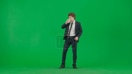 Foto de Moderno empresario creativo concepto de publicidad. Retrato de modelo masculino en traje en pantalla verde croma clave. Guapo joven hombre de negocios en traje de moda sosteniendo la taza de café y bailando - Imagen libre de derechos