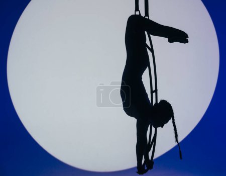 Téléchargez les photos : Silhouette de gymnaste féminin suspendu sur la lune en lumière bleue avec projecteur au centre. Acrobat femme démontre élément de performance sur la lune de l'air. Concept de gymnastique aérienne. - en image libre de droit