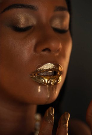 Téléchargez les photos : Portrait d'une jeune afro-américaine avec un collier en or autour du cou. L'or liquide est appliqué sur les lèvres des modèles et s'écoule dans les ruisseaux minces. Concept beauté. Gros plan - en image libre de droit