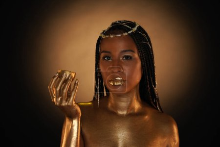 Photo for Portrait of an African-American woman with gold jewelry in her hair. The models arms, neck and shoulders are covered with liquid gold. The girl is holding a golden lemon in her hands. Close up. - Royalty Free Image