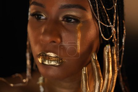 Photo for Portrait of an African-American woman with gold jewelry in her hair. The models eyes, lips and hands are covered with liquid gold. A golden tear is flowing down the girls cheek. Close up. - Royalty Free Image