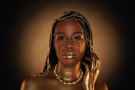 Foto de Retrato de una mujer afroamericana con joyas de oro en el pelo. Los modelos ojos, labios y manos están cubiertos de oro líquido. Una lágrima dorada fluye por la mejilla de las chicas. De cerca.. - Imagen libre de derechos