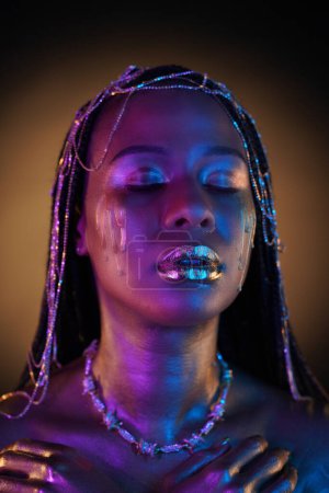 Foto de Mujer afroamericana con joyas de oro en el pelo. El cuerpo de los modelos cubiertos con oro líquido brilla en luz de neón violeta azul. Lágrimas doradas fluyen por las mejillas de las chicas. De cerca. - Imagen libre de derechos