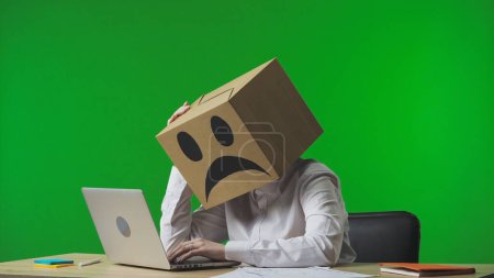 Téléchargez les photos : Femme dans une boîte en carton avec des émoticônes négatifs sur la tête sur fond vert studio. Un travailleur assis à un bureau, tapant sur un ordinateur portable, fatigué et frustré - en image libre de droit