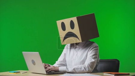 Photo for Woman in cardboard box with negative emoji on her head on green studio background. A worker sits at a desk, typing on a laptop. Business life and daily routine in the office - Royalty Free Image