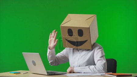 Foto de Mujer en caja de cartón con emoji sonriente en su cabeza sobre fondo verde estudio. Trabajadora hablando en videollamada usando laptop. Vida de negocios y rutina diaria en la oficina - Imagen libre de derechos