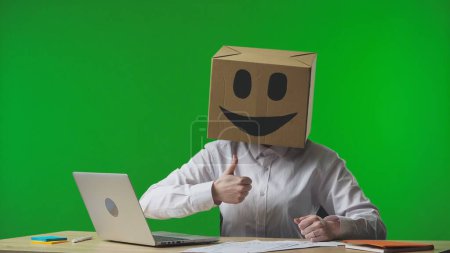 Téléchargez les photos : Femme dans une boîte en carton avec des émojis souriants sur la tête sur fond vert du studio. L'employé donne un pouce. Vie professionnelle. - en image libre de droit