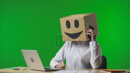 Téléchargez les photos : Femme dans une boîte en carton avec des émoticônes souriants sur la tête sur fond vert studio. Un employé qui parle sur un téléphone intelligent. La vie professionnelle et la routine quotidienne au bureau - en image libre de droit