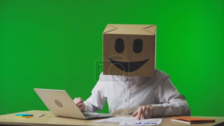 Photo for Woman in cardboard box with smiling emoji on her head on green background of studio. Employee checks data in documents and laptop and rejoices at her success. Business life. - Royalty Free Image