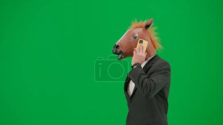 Téléchargez les photos : Homme en costume d'affaires avec masque de tête de cheval sur fond de studio vert. Homme d'affaires marchant et parlant sur smartphone. Concept de travail de bureau dur Demi tour - en image libre de droit