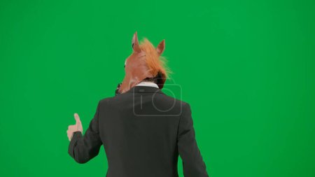 Téléchargez les photos : Homme en costume d'affaires avec masque de tête de cheval sur fond de studio vert. Homme d'affaires marchant, pointant les doigts dans la direction et montrant les pouces vers le haut geste. Concept de travail de bureau dur Vue arrière - en image libre de droit