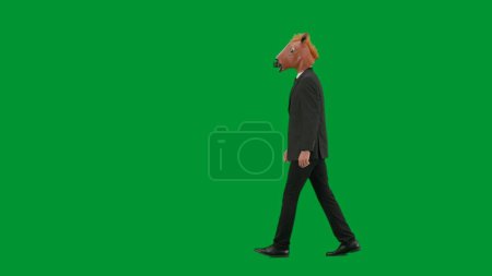 Téléchargez les photos : Homme en costume d'affaires avec masque de tête de cheval sur fond de studio vert. Homme d'affaires marchant à pied. Concept de travail de bureau dur Vue latérale - en image libre de droit