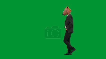 Téléchargez les photos : Homme en costume d'affaires avec masque de tête de cheval sur fond de studio vert. Homme d'affaires marchant et gardant les mains dans les poches du pantalon. Concept de travail de bureau dur Vue latérale - en image libre de droit