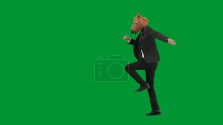 Téléchargez les photos : Homme en costume d'affaires avec masque de tête de cheval sur fond de studio vert. Homme d'affaires marchant, rebondissant et dansant. Concept de travail de bureau dur Vue latérale - en image libre de droit