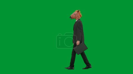 Téléchargez les photos : Homme en costume d'affaires avec masque de tête de cheval sur fond de studio vert. Homme d'affaires marchant avec une valise noire à la main. Concept de travail de bureau dur Vue latérale - en image libre de droit