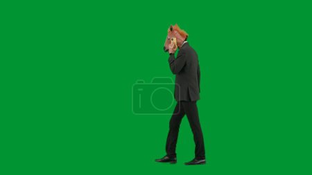 Téléchargez les photos : Homme en costume d'affaires avec masque de tête de cheval sur fond de studio vert. Homme d'affaires marchant et parlant sur smartphone. Concept de travail de bureau dur Vue latérale - en image libre de droit