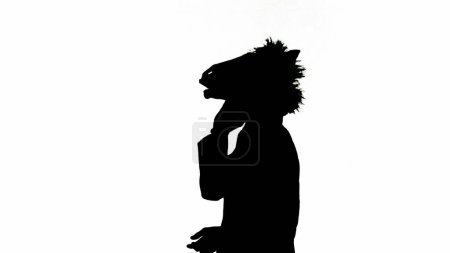 Foto de Silueta negra del hombre en traje de negocios con máscara de cabeza de caballo sobre fondo de estudio aislado blanco. Empresario caminando y hablando en smartphone. Concepto de trabajo duro de oficina - Imagen libre de derechos