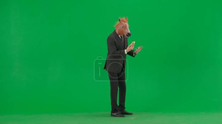 Téléchargez les photos : Un homme en costume d'affaires avec un masque de tête de cheval sur fond de studio vert. Un homme d'affaires parle sur un smartphone, se met en colère et crie dans le téléphone. Concept de travail de bureau dur - en image libre de droit