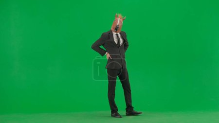Téléchargez les photos : Un homme en costume d'affaires avec un masque de tête de cheval sur fond de studio vert. Un homme d'affaires qui regarde la caméra. Concept de travail de bureau dur. - en image libre de droit