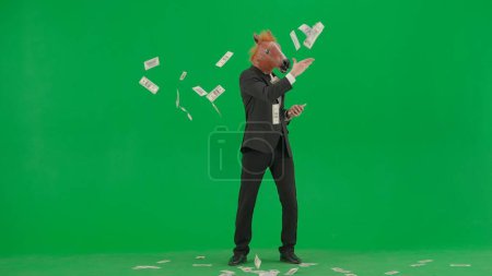 Foto de Un hombre con un traje de negocios con una máscara de caballo en un fondo de estudio verde. El hombre de negocios felizmente dispersa billetes. concepto de trabajo duro de oficina - Imagen libre de derechos