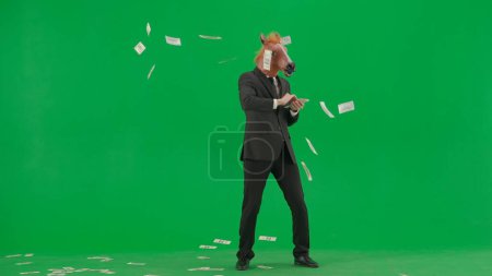 Téléchargez les photos : Un homme en costume d'affaires avec un masque de tête de cheval sur fond de studio vert. L'homme d'affaires disperse volontiers les billets. Concept de travail de bureau dur - en image libre de droit