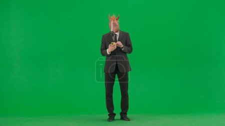 Téléchargez les photos : Un homme en costume d'affaires avec un masque de tête de cheval sur fond de studio vert. Un homme d'affaires regarde quelque chose sur son smartphone. Concept de travail de bureau dur - en image libre de droit