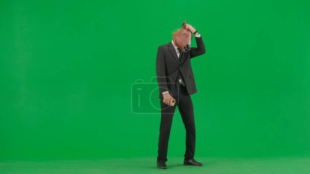 Téléchargez les photos : Un homme en costume d'affaires avec un masque de tête de cheval sur fond de studio vert. Un homme d'affaires regarde quelque chose sur son smartphone et est contrarié de perdre. Concept de travail de bureau dur - en image libre de droit
