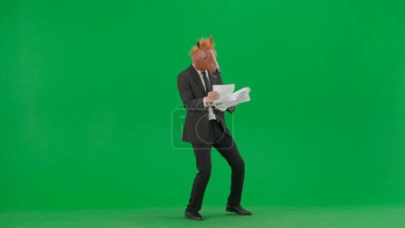 Téléchargez les photos : Un homme en costume d'affaires avec un masque de tête de cheval sur fond de studio vert. Un homme d'affaires regarde à travers des documentaires et danse joyeusement. Concept de travail de bureau dur - en image libre de droit
