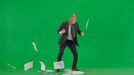 A man in a business suit with a horse head mask on a green studio background. A businessman looks through papers with charts, gets angry and scatters them. Hard office work concept