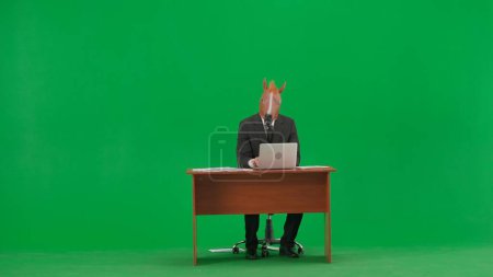 Téléchargez les photos : Homme en costume d'affaires avec masque de tête de cheval sur fond vert studio. Homme d'affaires assis au bureau et tapant sur le clavier de l'ordinateur portable. Concept de travail de bureau lourd - en image libre de droit