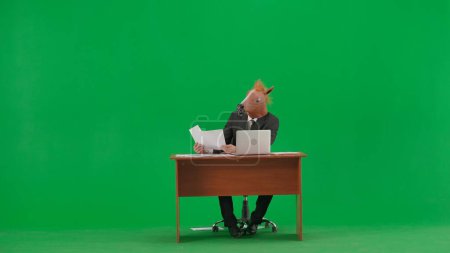 Téléchargez les photos : Homme en costume d'affaires avec masque de tête de cheval sur fond vert studio. Homme d'affaires assis au bureau regardant à travers les documents et travaillant à l'ordinateur portable. Concept de travail de bureau lourd - en image libre de droit
