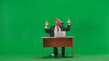 Téléchargez les photos : Homme en costume d'affaires avec masque de tête de cheval sur fond vert studio. Homme d'affaires assis au bureau les mains en l'air. Concept de travail de bureau lourd - en image libre de droit