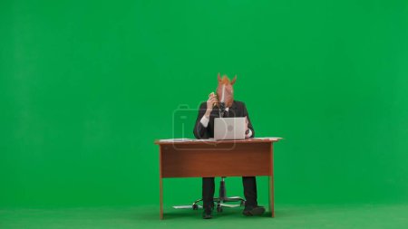 Téléchargez les photos : Homme en costume d'affaires avec masque de tête de cheval sur fond vert studio. Homme d'affaires assis au bureau et parlant sur smartphone. Concept de travail de bureau lourd - en image libre de droit