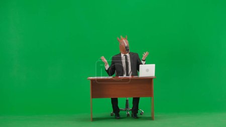 Foto de Hombre de traje de negocios con máscara de cabeza de caballo sobre fondo verde estudio. El hombre de negocios se sienta en el escritorio, dice algo y señala su mano hacia los lados. Concepto de trabajo pesado de oficina - Imagen libre de derechos