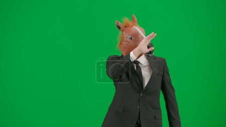 Téléchargez les photos : Homme en costume d'affaires avec masque de tête de cheval sur fond de studio vert. Homme d'affaires marchant et saluant. Concept de travail de bureau dur - en image libre de droit