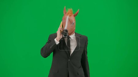 Foto de Hombre de traje de negocios con máscara de cabeza de caballo sobre fondo de estudio verde. Empresario caminando y hablando en smartphone. Concepto de trabajo duro de oficina - Imagen libre de derechos