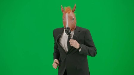 Téléchargez les photos : Homme en costume d'affaires avec masque de tête de cheval sur fond de studio vert. L'homme d'affaires court vite. Concept de travail de bureau dur - en image libre de droit