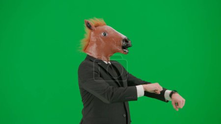 Téléchargez les photos : Un homme en costume d'affaires avec un masque de tête de cheval sur fond de studio vert. Concept de travail de bureau dur. - en image libre de droit