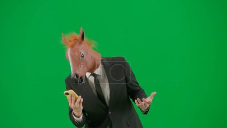 Téléchargez les photos : Un homme en costume d'affaires avec un masque de tête de cheval sur fond de studio vert. Un homme d'affaires regarde quelque chose sur son smartphone. Concept de travail de bureau dur - en image libre de droit