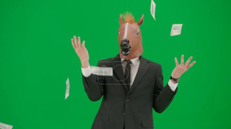 Téléchargez les photos : Un homme en costume d'affaires avec un masque de tête de cheval sur fond de studio vert. L'homme d'affaires disperse volontiers les billets. Concept de travail de bureau dur - en image libre de droit