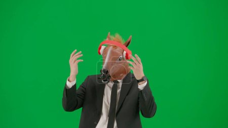 Téléchargez les photos : Un homme en costume d'affaires avec un masque de tête de cheval sur fond de studio vert. Un homme d'affaires écoute de la musique dans de grands écouteurs rouges et danse. Concept de travail de bureau dur - en image libre de droit