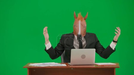 Téléchargez les photos : Homme en costume d'affaires avec masque de tête de cheval sur fond vert studio. Homme d'affaires assis au bureau tapant sur le clavier de l'ordinateur portable et très en colère. Concept de travail de bureau lourd - en image libre de droit