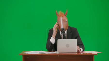 Photo for Man in business suit with horse head mask on studio green background. Businessman sitting at desk and talking on smartphone. Heavy office work concept - Royalty Free Image