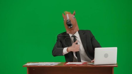 Téléchargez les photos : Homme en costume d'affaires avec masque de tête de cheval sur fond vert studio. Homme d'affaires assis au bureau et montrant les pouces vers le haut geste. Concept de travail de bureau dur - en image libre de droit