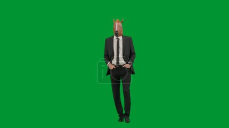 Téléchargez les photos : Homme en costume d'affaires avec masque de tête de cheval sur fond de studio vert. Homme d'affaires marchant et gardant les mains dans les poches du pantalon. Concept de travail de bureau dur - en image libre de droit