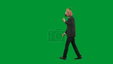 Téléchargez les photos : Homme en costume d'affaires avec masque de tête de cheval sur fond de studio vert. Homme d'affaires marchant, pointant du doigt dans la direction. Concept de travail de bureau dur Vue latérale - en image libre de droit
