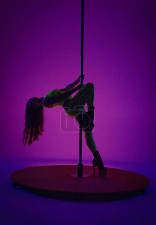 Photo for A young woman in a yellow neon mesh bodysuit posing near a pylon pole. Dancer on high heels in studio on pink background with blue neon light - Royalty Free Image