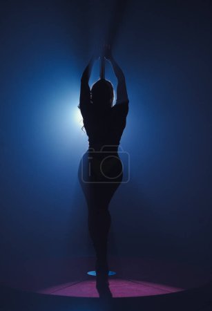 Foto de Silueta oscura de mujer flexible y plástica bailando sobre asta. Bailarina con pelo largo posando sobre pilón en estudio oscuro contra proyector brillante - Imagen libre de derechos